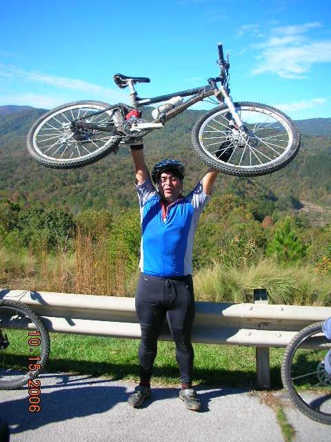 A person wearing a blue cycling jersey and black pants is standing next to a bike, holding it above their head in celebration. They are outdoors, with a scenic backdrop of rolling hills and trees under a clear blue sky. Tanasi Trail System mountain bike trail.