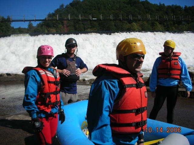 Four individuals in colorful life jackets and helmets stand near a blue inflatable raft, with a large waterfall in the background. The group appears to be preparing for a whitewater rafting adventure. The photo is taken on a sunny day, with trees and a bridge visible in the scenery. Tanasi Trail System mountain bike trail.