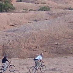 Two people riding bicycles on a rocky, desert landscape with gentle hills in the background. Slickrock mountain bike trail.