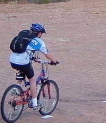 A person riding a mountain bike on a gravel path, wearing a blue helmet and a cycling jersey with shorts. They have a backpack secured on their back. Slickrock mountain bike trail.