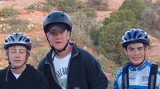 Three young males wearing helmets pose together outdoors amidst a natural landscape. Two are dressed in casual attire, while one is in a cycling jersey. The background features rocky terrain and greenery. Slickrock mountain bike trail.