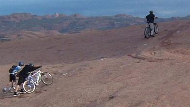 Two cyclists are walking their bikes up a rocky slope while a third cyclist rides ahead on a dirt path in a rugged outdoor landscape with distant hills. Slickrock mountain bike trail.