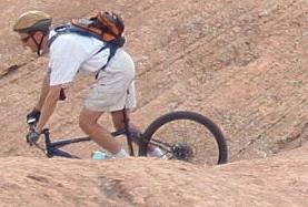 A cyclist wearing a helmet and a backpack rides down a rocky, uneven terrain on a mountain bike. Slickrock mountain bike trail.