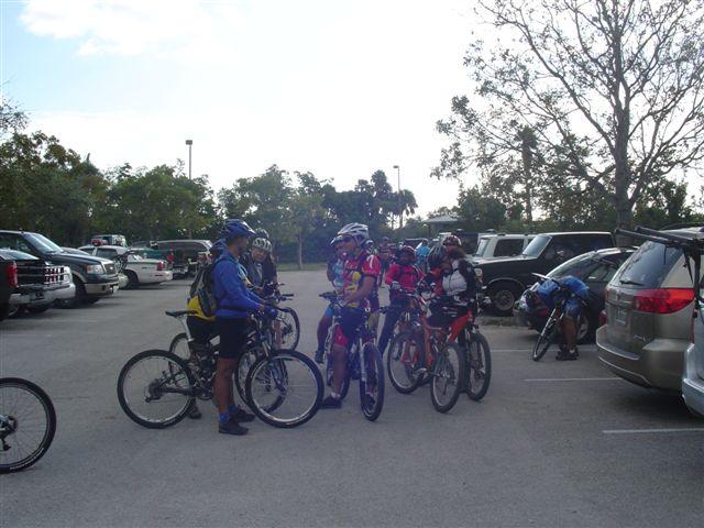 A group of mountain bikers gathered in a parking lot, wearing helmets and cycling gear, with their bicycles nearby. Several vehicles are parked in the background, and trees can be seen in the distance under a partly cloudy sky. Oleta River State Park mountain bike trail.