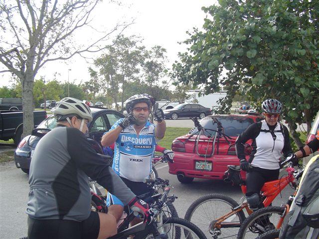 A group of cyclists in a parking area, preparing for a ride. One cyclist in a blue and white jersey gestures while discussing plans with others. Nearby, trees and parked cars are visible, creating a casual outdoor setting. Oleta River State Park mountain bike trail.