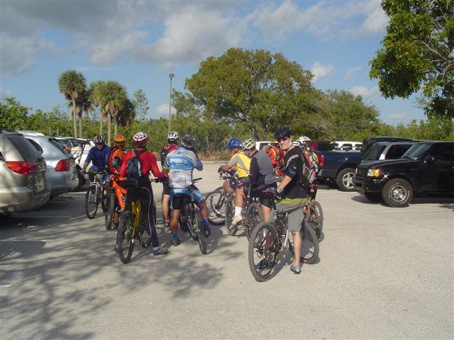 A group of cyclists in colorful jerseys and helmets gathered near their bikes in a parking lot, with cars parked in the background and trees under a partly cloudy sky. Oleta River State Park mountain bike trail.
