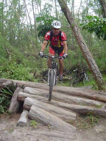 A cyclist dressed in a red and yellow jersey rides over a series of wooden logs in a forested area, surrounded by green foliage. A second bike is visible in the background, partially obscured by trees. Oleta River State Park mountain bike trail.