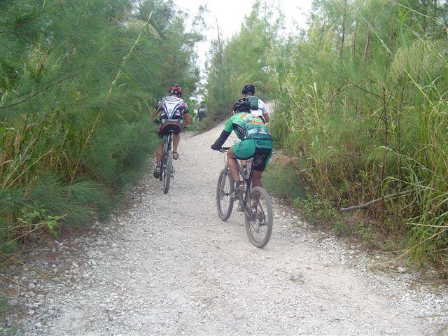 Two mountain bikers riding on a gravel path surrounded by greenery and trees, heading away from the camera. The cyclists are wearing helmets and sports attire, and the trail winds through a natural landscape. Oleta River State Park mountain bike trail.