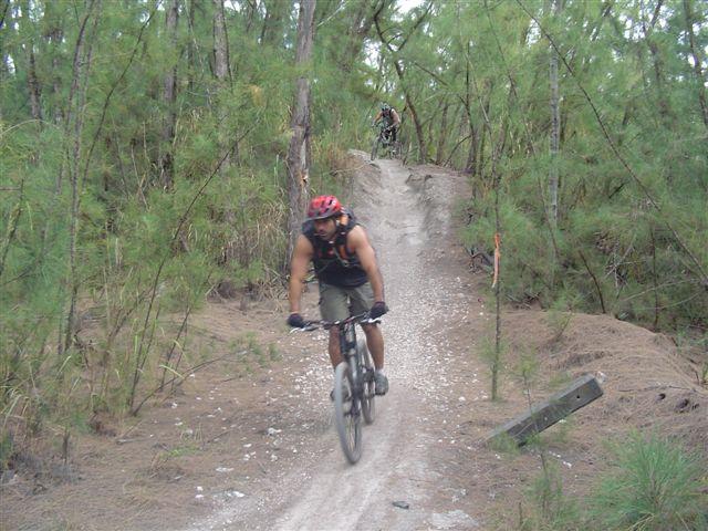 Two mountain bikers riding on a dirt trail surrounded by trees. The foreground features a biker wearing a red helmet and a sleeveless shirt, maneuvering down a slope. A second biker can be seen in the background, ascending a hill on the same trail. The scene captures a natural, rugged outdoor environment. Oleta River State Park mountain bike trail.
