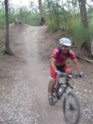 A mountain biker in red cycling gear rides down a dirt path, while another person is seen in the background preparing to descend from a small hill in a wooded area. The scene conveys an active outdoor environment, surrounded by trees and gravel trails. Oleta River State Park mountain bike trail.
