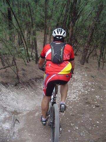 A cyclist wearing a red and yellow jersey and a helmet rides a mountain bike along a dirt path surrounded by trees. The view is from behind, showing the cyclist's back and the rugged terrain ahead. Oleta River State Park mountain bike trail.