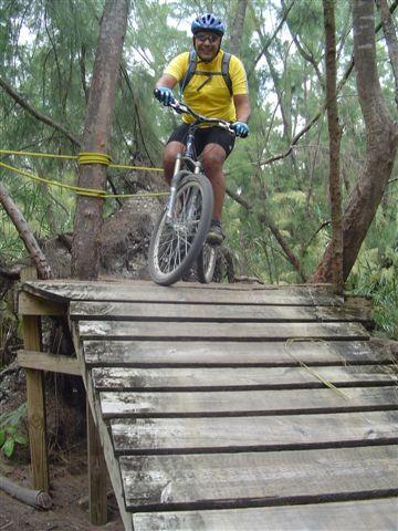 A mountain biker in a yellow shirt and helmet rides over a wooden bridge in a forested area, surrounded by trees. Oleta River State Park mountain bike trail.