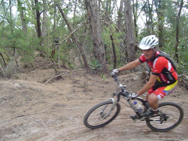 A mountain biker riding along a dirt trail through a wooded area, wearing a red and yellow cycling outfit and a helmet. The cyclist is in motion, with trees and underbrush visible in the background. Oleta River State Park mountain bike trail.