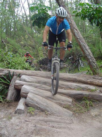 A mountain biker in a blue jersey and helmet navigates over a series of wooden logs on a dirt trail surrounded by greenery. Other bicycles are visible in the background. Oleta River State Park mountain bike trail.