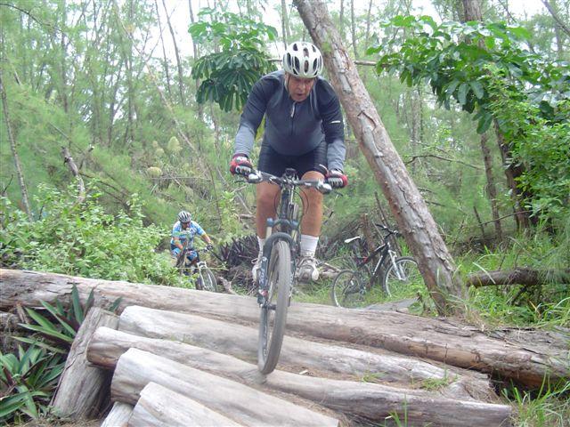 A mountain biker navigates over a log bridge in a dense forest, while another cyclist follows in the background. The scene captures the thrill of biking in nature, surrounded by greenery and trees. Oleta River State Park mountain bike trail.