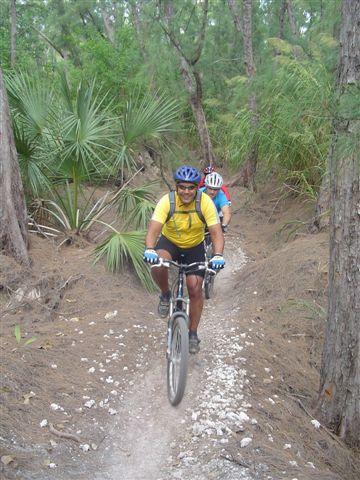 Two mountain bikers navigating a dirt trail through a wooded area with palm trees. The first rider, wearing a yellow shirt and blue helmet, is smiling as he rides, while a second rider in a red and blue outfit follows closely behind. Oleta River State Park mountain bike trail.