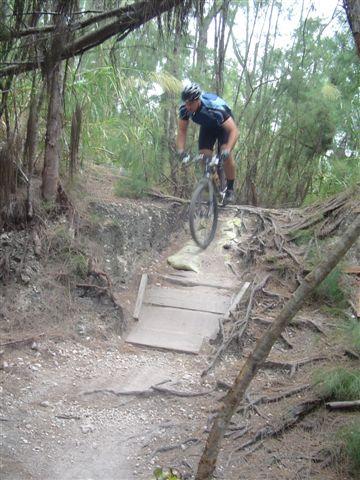A mountain biker wearing a helmet and blue jersey performs a jump on a dirt trail surrounded by trees and greenery. The biker is airborne over a small wooden ramp, with the bike positioned mid-air and terrain visible below. Oleta River State Park mountain bike trail.