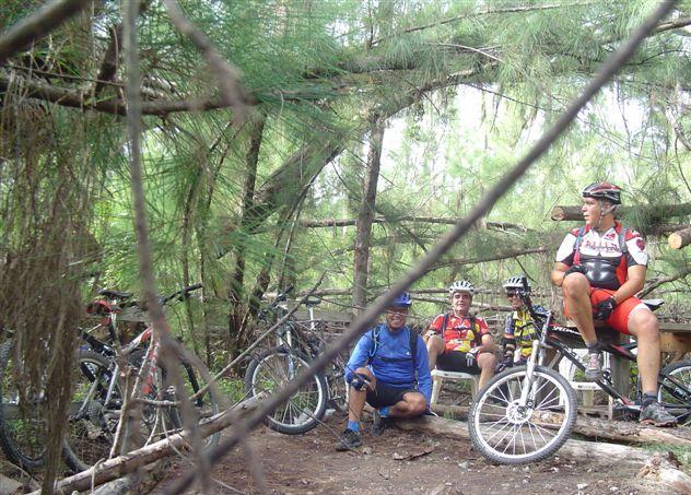Three mountain bikers resting in a forested area, surrounded by trees. Two bikers are seated on a bench made of logs, while one is perched on his bike. Several mountain bikes are visible nearby, and the scene conveys a sense of camaraderie and outdoor adventure. Oleta River State Park mountain bike trail.