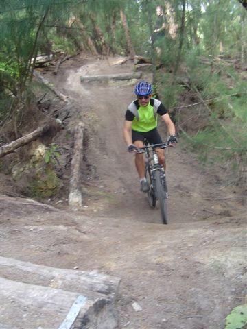 A mountain biker riding on a dirt trail surrounded by trees, wearing a helmet and a bright green shirt. The path is slightly downhill and includes obstacles like wooden logs. Oleta River State Park mountain bike trail.