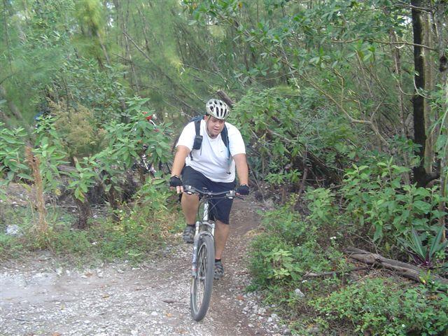 A person riding a mountain bike on a narrow dirt trail surrounded by lush greenery and trees. The rider is wearing a helmet and a backpack, navigating the bike path through a vibrant natural setting. Oleta River State Park mountain bike trail.