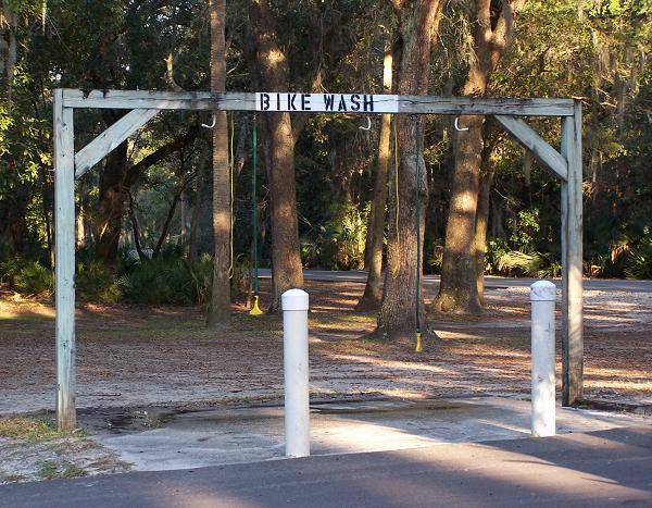 A wooden structure labeled "BIKE WASH" with two hanging hooks and white cylindrical posts, set in a natural outdoor environment with trees and gravel. Kathryn Abby Hanna Park mountain bike trail.