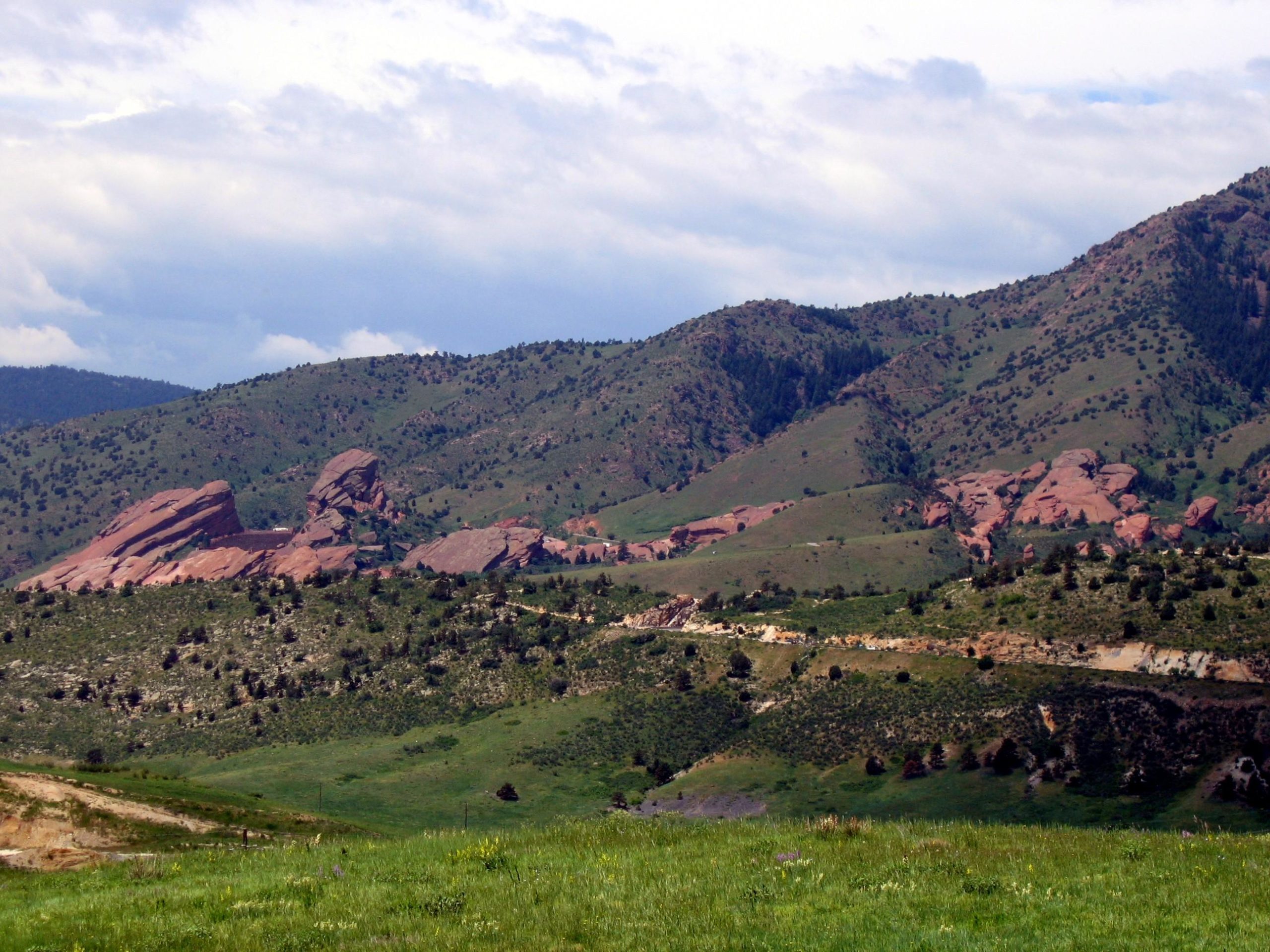 A scenic view of rolling green hills and reddish rock formations under a partly cloudy sky. The landscape features lush vegetation and scattered trees in the foreground, with a backdrop of mountains. Green Mountain mountain bike trail.