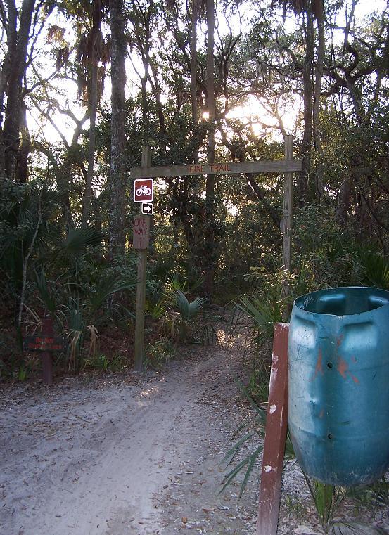 A dirt path leads into a wooded area, marked by a wooden sign that reads "BIKE TRAIL." A bicycle symbol indicates that the trail is intended for biking, and a smaller sign denotes that it is "ONE WAY." In the foreground, there is a green, cylindrical container beside the trail. The scene is surrounded by lush trees and foliage, with sunlight filtering through the branches. Kathryn Abby Hanna Park mountain bike trail.