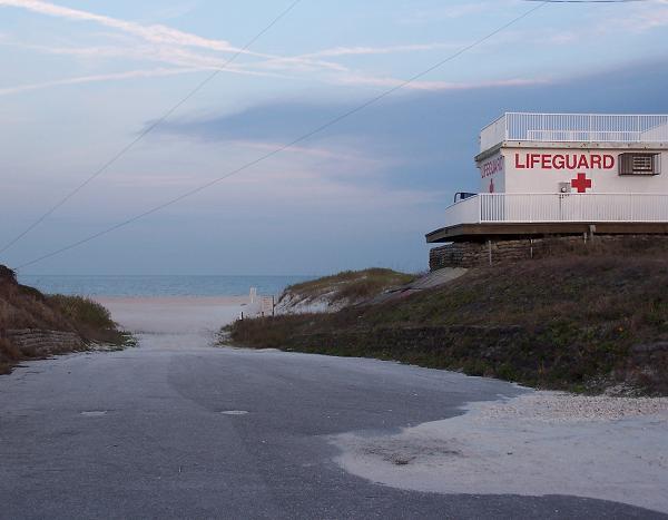 A quiet beach entrance with a road leading towards the ocean. On the right, there is a lifeguard station with a prominent "LIFEGUARD" sign. The scene features soft sandy beach and grasses on either side, under a partly cloudy sky. Kathryn Abby Hanna Park mountain bike trail.