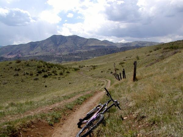 A dirt trail winding through grassy hills, with a view of mountains in the background under a cloudy sky. Two bicycles are leaning against the trail, suggesting a scenic spot for outdoor enthusiasts. Green Mountain mountain bike trail.