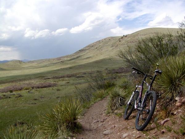 A scenic landscape featuring a dirt trail winding through a green, grassy area, with a mountain in the background. A mountain bike is leaning against a bush on the side of the path, under a partly cloudy sky. Green Mountain mountain bike trail.