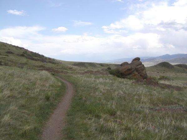 A narrow dirt path winds through a grassy landscape, with rolling hills and a large rock on one side. The sky is partly cloudy, with hints of blue peeking through. The scene evokes a sense of tranquility and openness in nature. Green Mountain mountain bike trail.