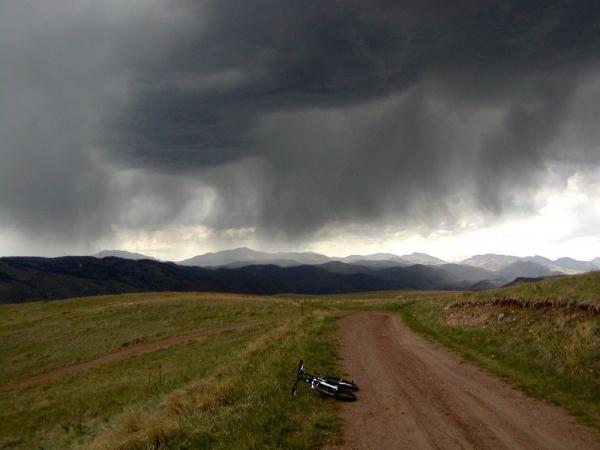 A dirt path stretches through a grassy landscape, leading towards distant mountains under a dark, stormy sky filled with heavy clouds. A bicycle lies on its side near the edge of the path, suggesting an abrupt stop or a quick departure. Green Mountain mountain bike trail.