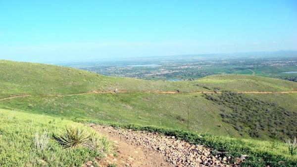 A panoramic view of rolling green hills under a clear blue sky, with a winding dirt path leading through the landscape. In the distance, small towns and roads are visible against the horizon, showcasing a tranquil rural setting. Green Mountain mountain bike trail.