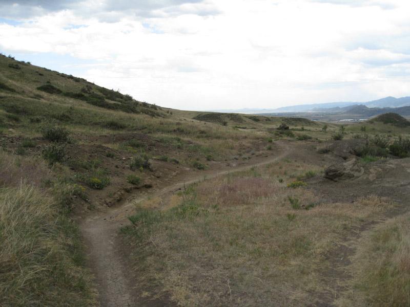 A winding dirt path through a grassy landscape, surrounded by rolling hills and sparse vegetation under a cloudy sky. Green Mountain mountain bike trail.