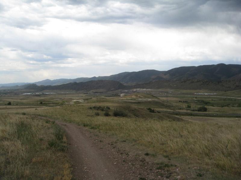 A dirt path winds through grassy terrain, leading towards a distant view of mountains under a partly cloudy sky. The landscape is expansive, showcasing rolling hills and open fields, with a hint of development visible in the background. Green Mountain mountain bike trail.