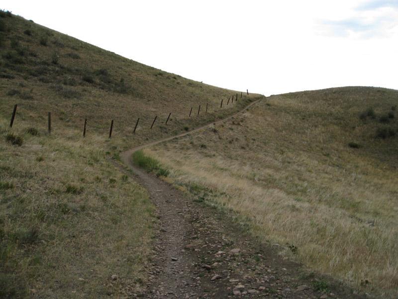 A winding dirt path leads through grassy hills, bordered by a sparse barbed-wire fence. The trail ascends into the distance, under a cloudy sky. Green Mountain mountain bike trail.