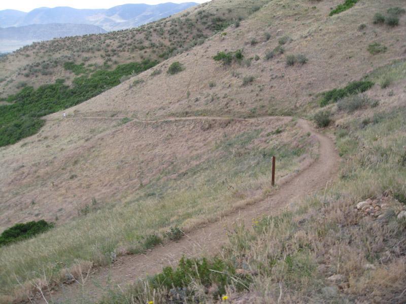 A winding dirt path through a grassy hillside, with sparse vegetation and rolling mountains in the background under a cloudy sky. Green Mountain mountain bike trail.