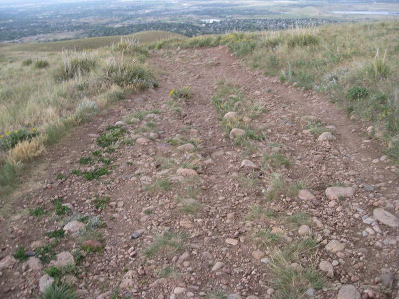 A dirt trail winding through grassy terrain with scattered rocks and sparse vegetation, leading towards a distant landscape. Green Mountain mountain bike trail.