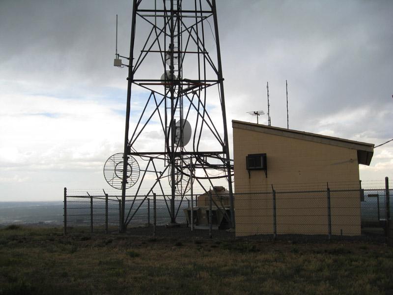A tall radio tower with multiple antennas stands next to a small building, both located in a fenced area on a grassy hilltop. The sky is overcast, with dark clouds visible in the background. Green Mountain mountain bike trail.