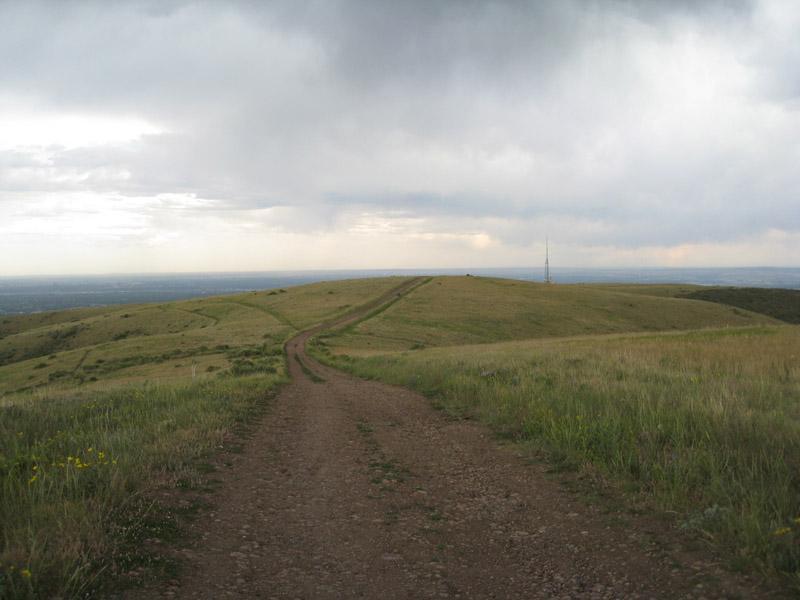 A dirt path winding through grassy hills under a cloudy sky, leading towards a small hilltop with a communication tower in the distance. Green Mountain mountain bike trail.