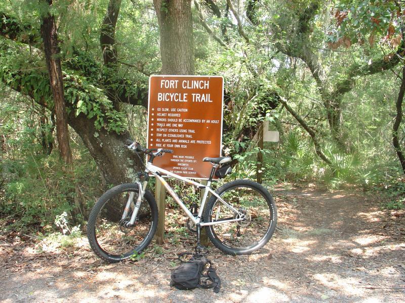 A mountain bike rests beside a sign for the Fort Clinch Bicycle Trail, surrounded by lush greenery. The sign provides trail guidelines, including safety precautions and rules for riders. In the background, a dirt path leads into the densely wooded area. Ft. Clinch State Park mountain bike trail.