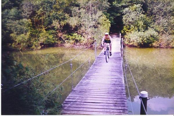 A person riding a mountain bike across a narrow, wooden suspension bridge over a calm body of water, surrounded by lush greenery and trees on either side. Hard Rock mountain bike trail.