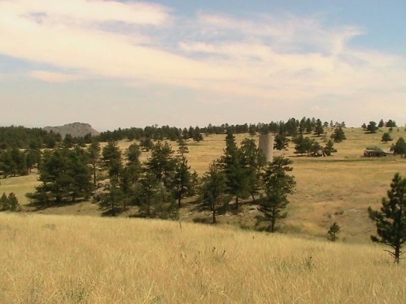 A scenic landscape featuring rolling hills covered with tall grass and clusters of evergreen trees, with a distant rocky outcrop visible on the left. In the middle ground, there is a silvery cylindrical structure and a rustic building nestled among the trees, under a partly cloudy blue sky. Hall Ranch mountain bike trail.