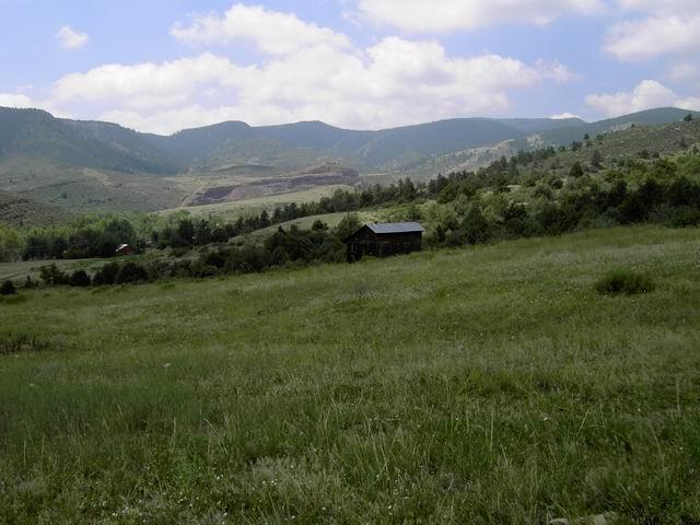 A serene landscape featuring a vast green field with scattered shrubs and a small wooden cabin. In the background, rolling hills and mountains are visible under a partly cloudy sky. The scene evokes a peaceful, rural atmosphere. Hall Ranch mountain bike trail.