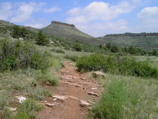 A dirt trail winding through a grassy landscape, flanked by shrubs and small trees, leads towards a prominent rocky formation or mesa in the background under a partly cloudy sky. Hall Ranch mountain bike trail.