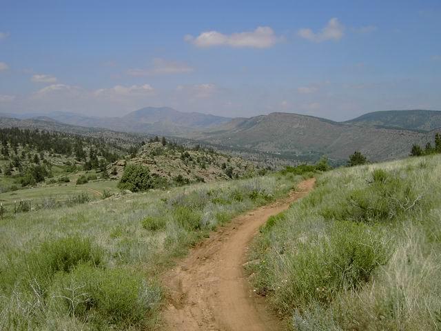 A dirt path winds through green grass and shrubs, leading toward rolling hills and mountains under a partly cloudy sky. Hall Ranch mountain bike trail.
