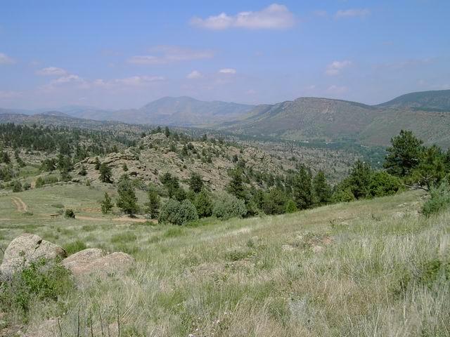 A scenic view of rolling hills and rocky terrain, dotted with trees and shrubs, under a blue sky with fluffy white clouds. The landscape gradually ascends towards distant mountains, creating a tranquil and natural setting. Hall Ranch mountain bike trail.