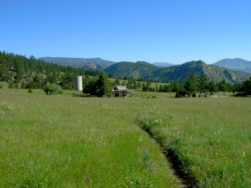 A scenic view of a grassy field leading to an old, rustic cabin and a grain silo, surrounded by trees and rolling mountains under a clear blue sky. Hall Ranch mountain bike trail.