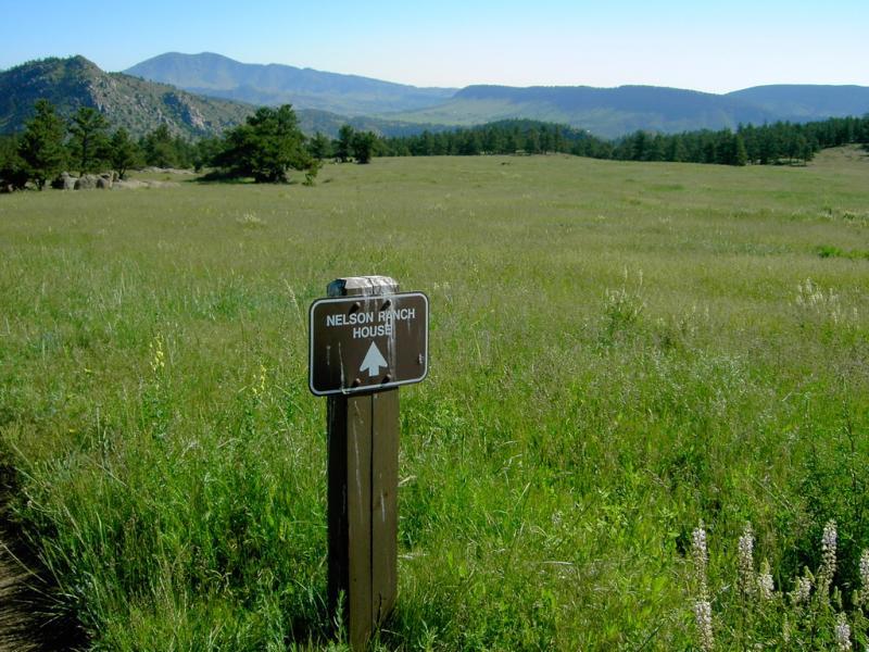 Sign indicating direction to Nelson Ranch House amidst a vast, grassy landscape with mountains in the background. The scene features a clear blue sky and scattered trees in the distance. Hall Ranch mountain bike trail.