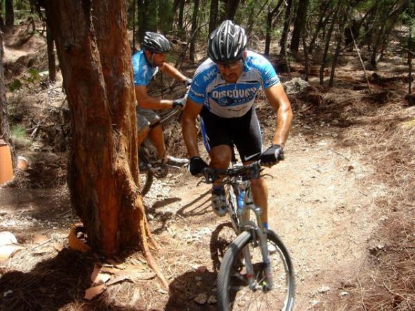 Two mountain bikers riding on a narrow trail through a forest. One biker is in the foreground, wearing a blue and white jersey, and is navigating around a tree, while the second biker is visible in the background slightly blurred and also wearing a jersey. The ground is covered in pine needles and dirt, with trees surrounding the path. Oleta River State Park mountain bike trail.